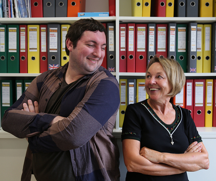 Christian Dobnik and Anna Maria Moisi laughing heartily, each with arms crossed, side by side, in front of a wall covered with files in the office at TU Graz.