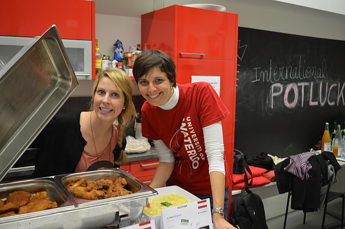 2 junge Frauen, Mitarbeiterinnen des TU Graz-Welcome Centers, öffnen bei einem Kochevent für internationale Studierende gut gelaunt einen Warmhaltebehälter mit Schnitzeln.