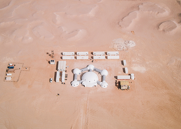 Aerial photo of the station, one sees desert sand and the station in the center surrounded by white containers.