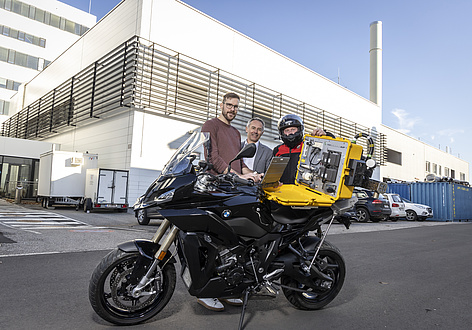 Three men are standing behind a motorbike with a yellow case containing a measuring device.