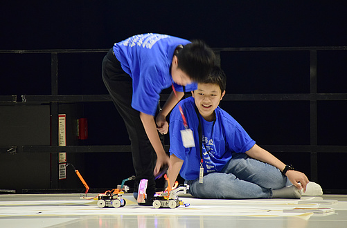 Two teenagers observe a small robot travelling along the floor.
