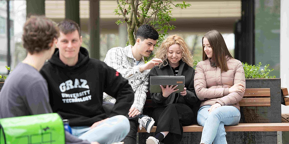 Students – three young men and two young women – on benches in front of a university building.