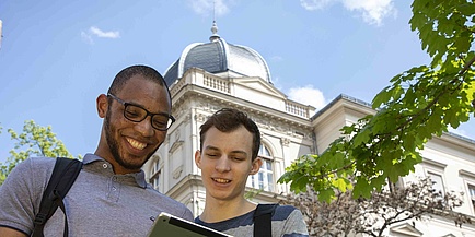 Zwei männliche Studenten unterschiedlicher ethnischer Herkunft blicken auf ein Tablet. Im Hintergrund ist ein historisches Universitätsgebäude.