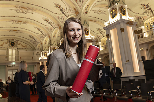 A woman smiles at the camera and holds up a red tube with his doctorate certificate