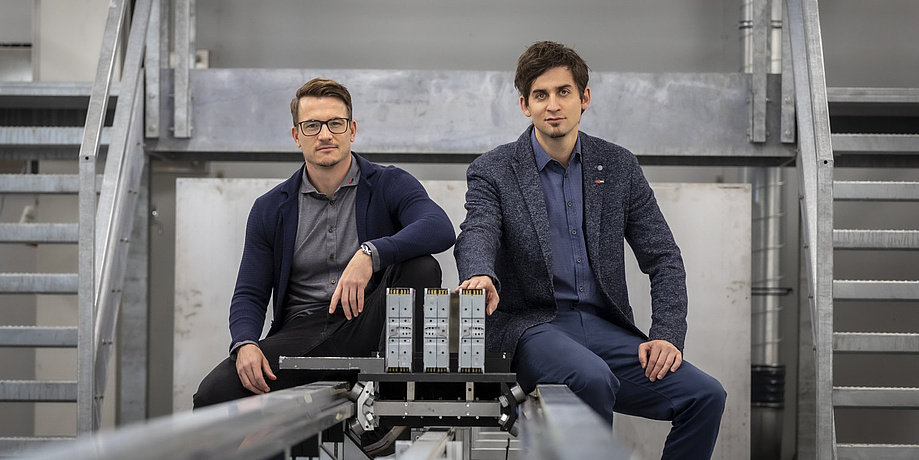 Two men are sitting on the rails of a test bench and are looking into the camera.