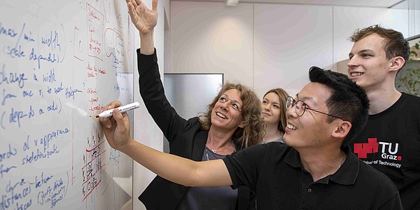 Drei Studierende unterschiedlicher Herkunft und eine Professorin unterhalten sich angeregt über Formeln am Whiteboard.