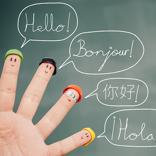 Four fingers in front of a board, which displays faces which wear different sorts of hats. The board says „Hello“ in different languages. Photo source: lemontreeimages - fotolia.com
