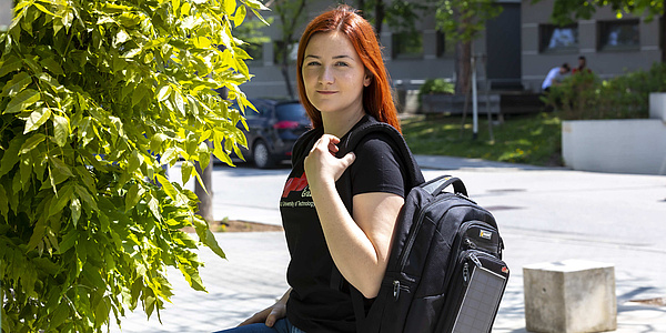 Young woman with a rucksack is sitting in front of trees and a building in the background.