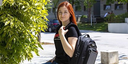 Eine Studentin mit Rucksack steht am Campus, neben ihr die Blätter einer Pflanze.