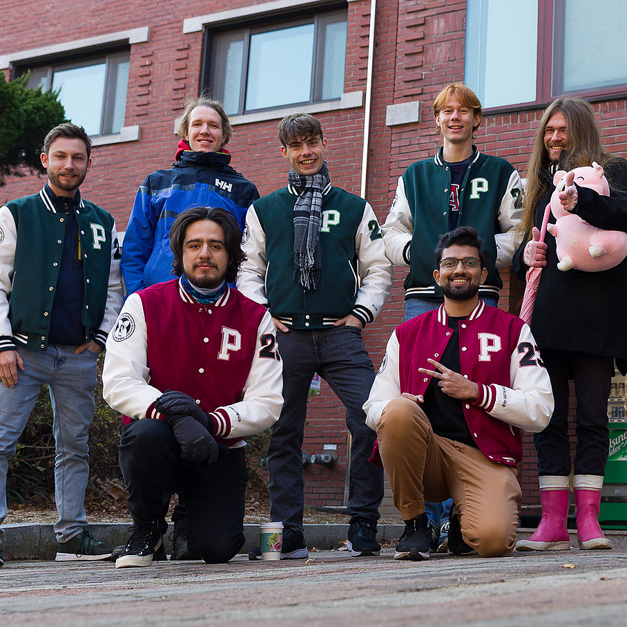 Eine Gruppe internationaler Studierender in Collegejacken stehen vor einem Universitätsgebäude.