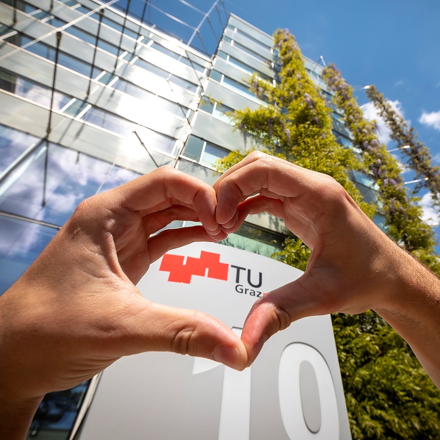 Two hands form a heart in front of a large building and a sign with the TU Graz logo.