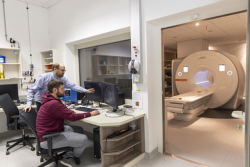 Two men are standing at a desk looking at a computer screen. In the background is an MRI machine.