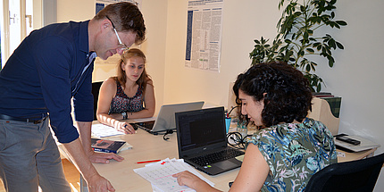 Two woman are working at a desk. A man stands besides them.