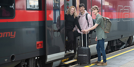 Drei Studierende mit Handgepäck und Koffern steigen am Bahnhof in einen Zug.