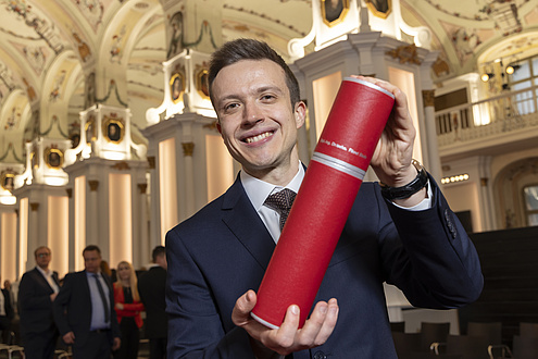 A man smiles at the camera and holds up a red tube with his doctorate certificate