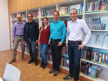 Five people are standing next to each other in front of a bookshelf.