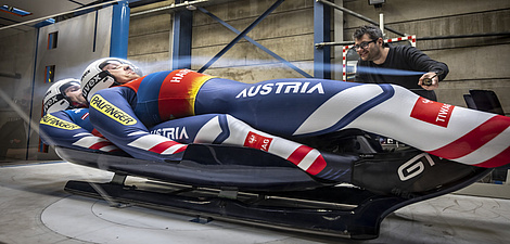 Two athletes are lying on a sled in a wind tunnel.