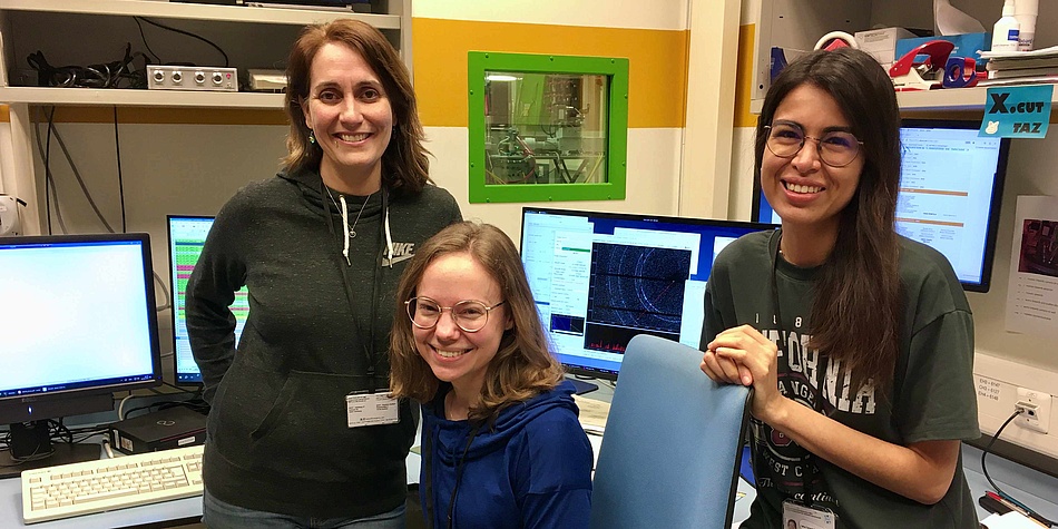 Drei Frauen in einem Labor. Dahinter sind Bildschirme und diverse Messsysteme zu sehen. 