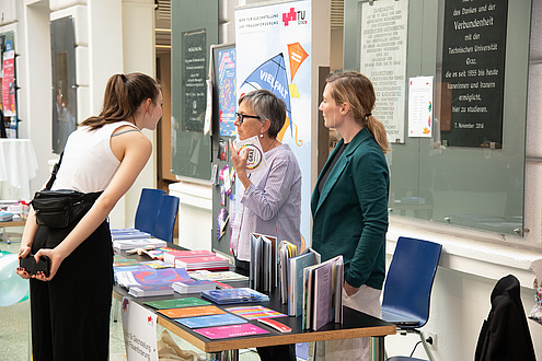 Two women are standing behind a row of tables with books and flyers in different colors. They are talking to a third woman standing on the other side of the row of tables.