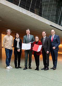 Sechs Personen stehen beieinander und schauen in die Kamera. Die beiden Personen in der Mitte halten Mappen in der Hand.