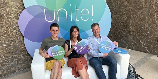 Three students holding up colourful signs representing the University Alliance "Unite!"