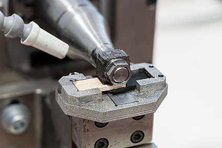 A machine presses from above onto a narrow wooden part that lies on a plastic part.