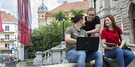 Ein junger Mann mit aufgeklapptem Laptop am Schoß und eine junge Frau sitzen auf einer Steinmauer vor einem historischen Gebäude. Ein zweiter junger Mann steht hinter ihnen. Alle drei sind im Gespräch.