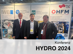 Drei Personen vor einem Stand der TU Graz bei der Konferenz HYDRO 2024, mit Bannern der Institute IWB, IEE und HFM im Hintergrund.