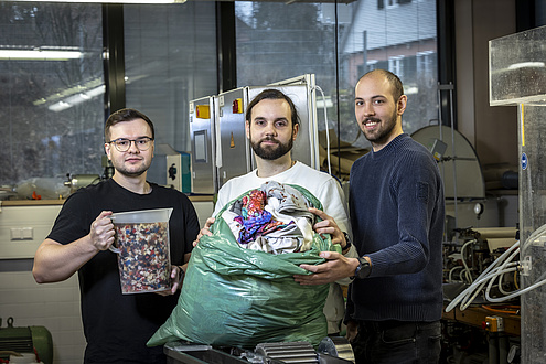 Three men are standing next to each other in a laboratory looking into the camera. The man on the left is holding a transparent jug in which small pieces of textile are floating. The man in the centre is holding a bag of old clothes.