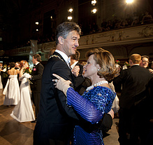 A man in a black suit and a woman in a blue ball gown dance. 