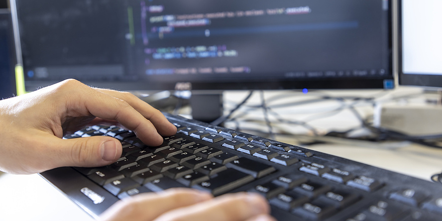 Hands are typing on a computer keyboard. A computer monitor with code on it is blurred in the background.