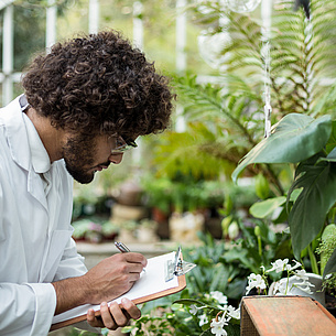 A man wearing a lab coat and safety glasses is standing between plants.