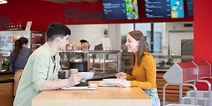 A young man and a young woman with plates in front of them sit opposite each other at a table in a canteen.