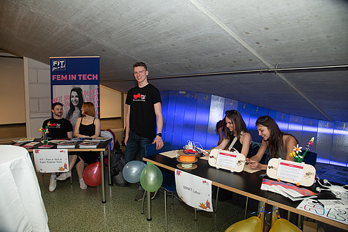 Several people are sitting at tables with information material on them. Colorful balloons are hanging from the tables. One person is standing between the tables.
