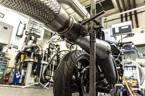 The rear of a motorbike photographed from diagonally below. The exhaust is connected to a pipe for measuring the exhaust gases.