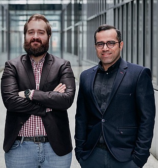 Two men smile at the camera. The man on the right is wearing a dark suit, the man on the left a leather jacket and a chequered shirt.