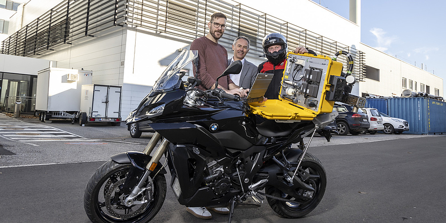 Three men are standing behind a motorbike with a yellow case containing a measuring device.