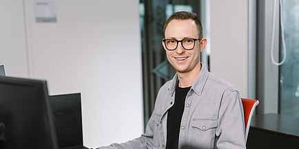 Ein junger Mann mit grauem Hemd sitzt an seinem Schreibtisch und blickt in die Kamera.