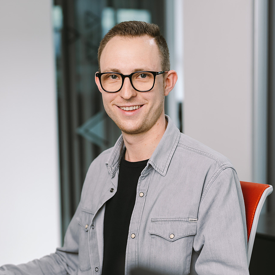 Ein junger Mann mit grauem Hemd sitzt an seinem Schreibtisch und blickt in die Kamera.