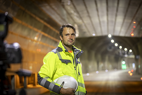 A man with a bright yellow jacket and a safety helmet under his arm is standing in a tunnel.