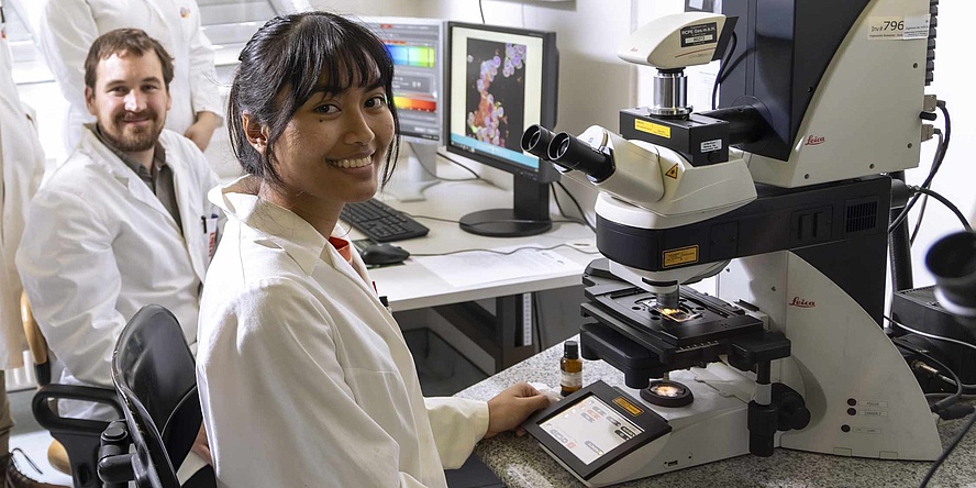 A woman in a white lab coat sits in front of a microscope, behind her sits a man with dark hair and a beard. 