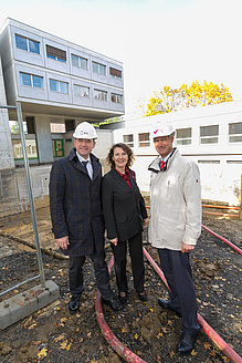Two men and a woman are standing on a construction site. The two men are wearing a white helmet. All three look into the camera.