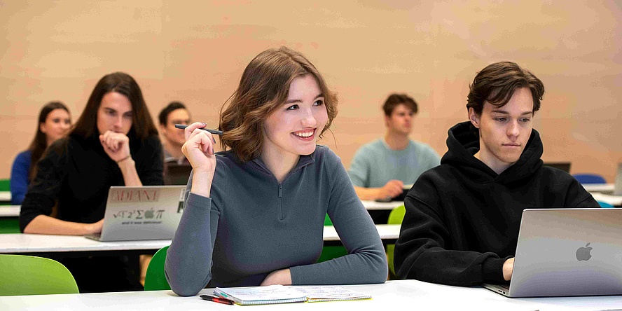 Weibliche und männliche Studierende mit aufgeklappten Laptops oder mit Blöcken vor sich am Tisch in einem Seminarraum.