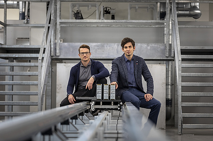 Two men are sitting on the rails of a test bench and looking into the camera.