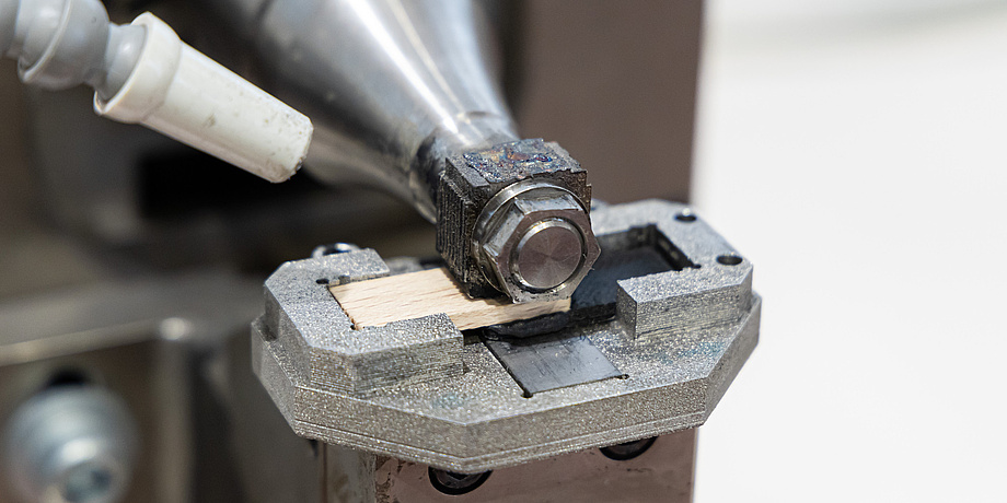 A machine presses from above onto a narrow wooden part that lies on a plastic part.