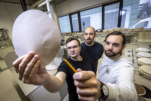 Three men are standing together looking at a round piece of paper that one of them is holding up.