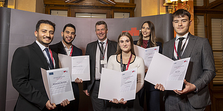 Six people, four men and two women, smile into the camera. Three of the men and the two women are holding certificates recognising them as scholarship recipients.