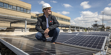 Ein Mann in schwarzer Jacke und mit weißem Schutzhelm inmitten von Solarpaneelen.