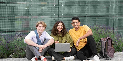 Eine Studentin mit Laptop und zwei Studenten unterschiedlicher Hautfarbe sitzen am Campusgelände im Schneidersitz.