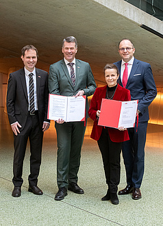 Four people stand together and look into the camera. The two people in the centre are holding folders.